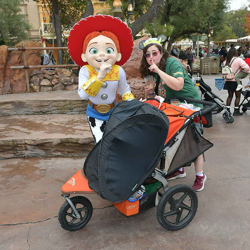 Person in a Jessie costume from Toy Story pushing a stroller with a child in a park setting.